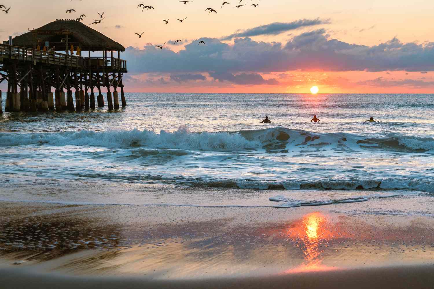 A group of surfers and a pier in the ocean at sunset.